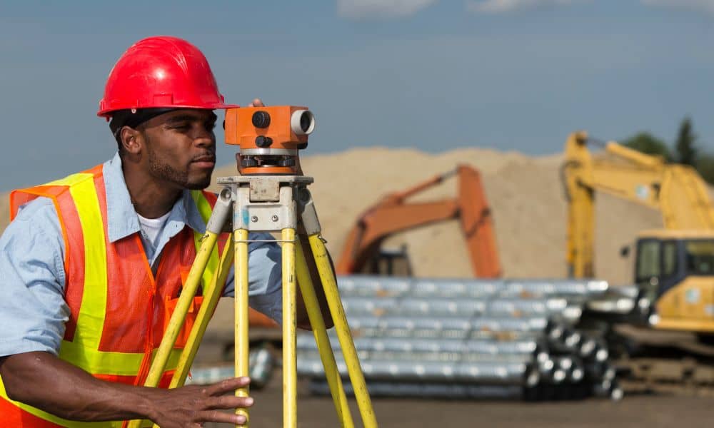Construction staking survey being performed by a land surveyor in safety gear at a construction site in Oklahoma City with heavy machinery in the background

