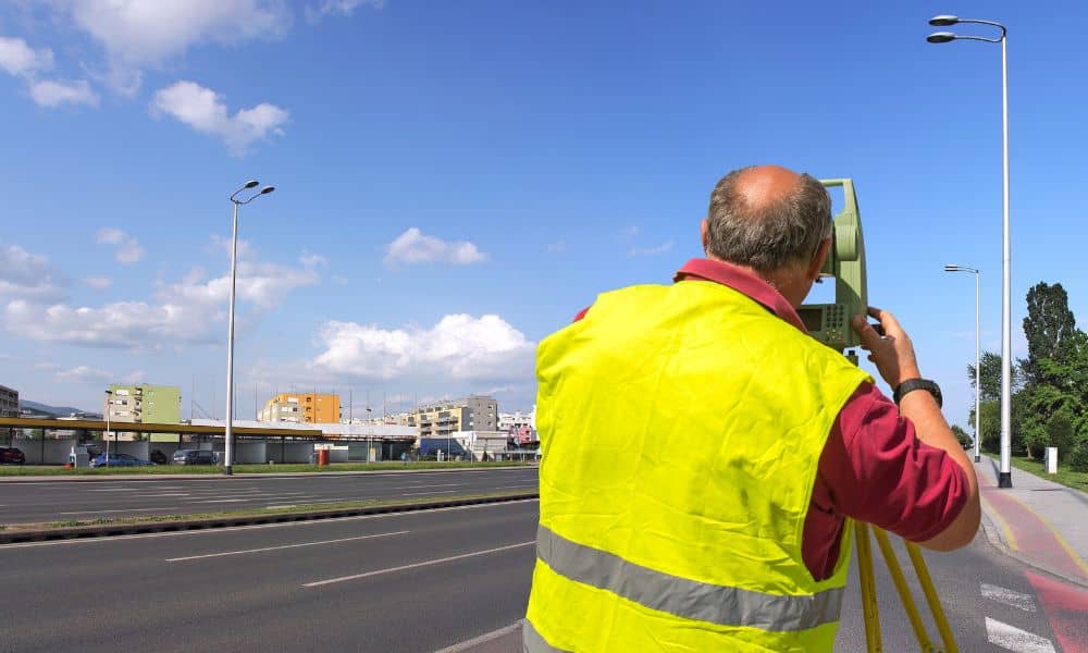 Surveyor using total station beside a city roadway during an ALTA Land Title Survey for infrastructure development
